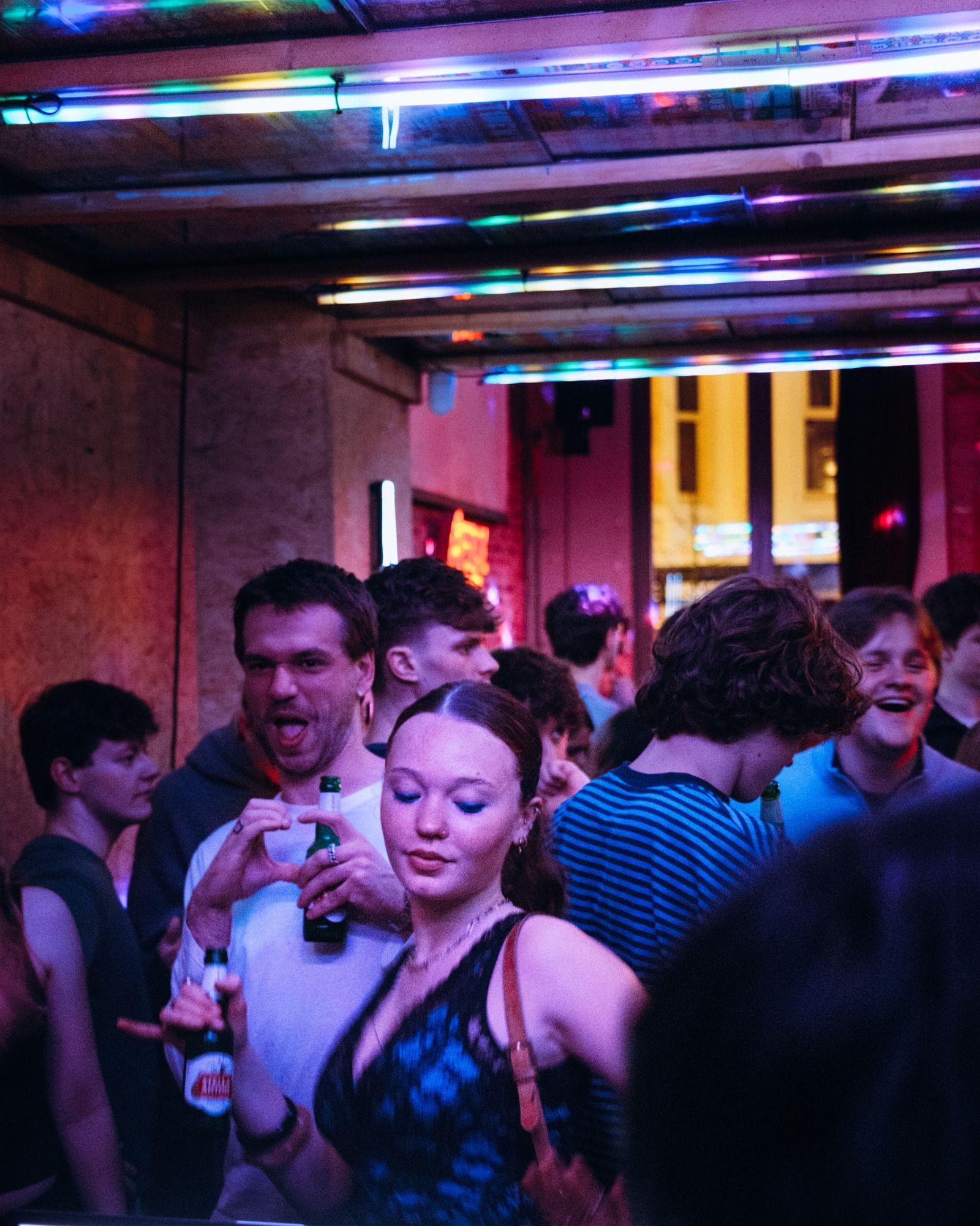 Mensen dansen met een flesje in de hand onder roze en blauwe neonbuizen.