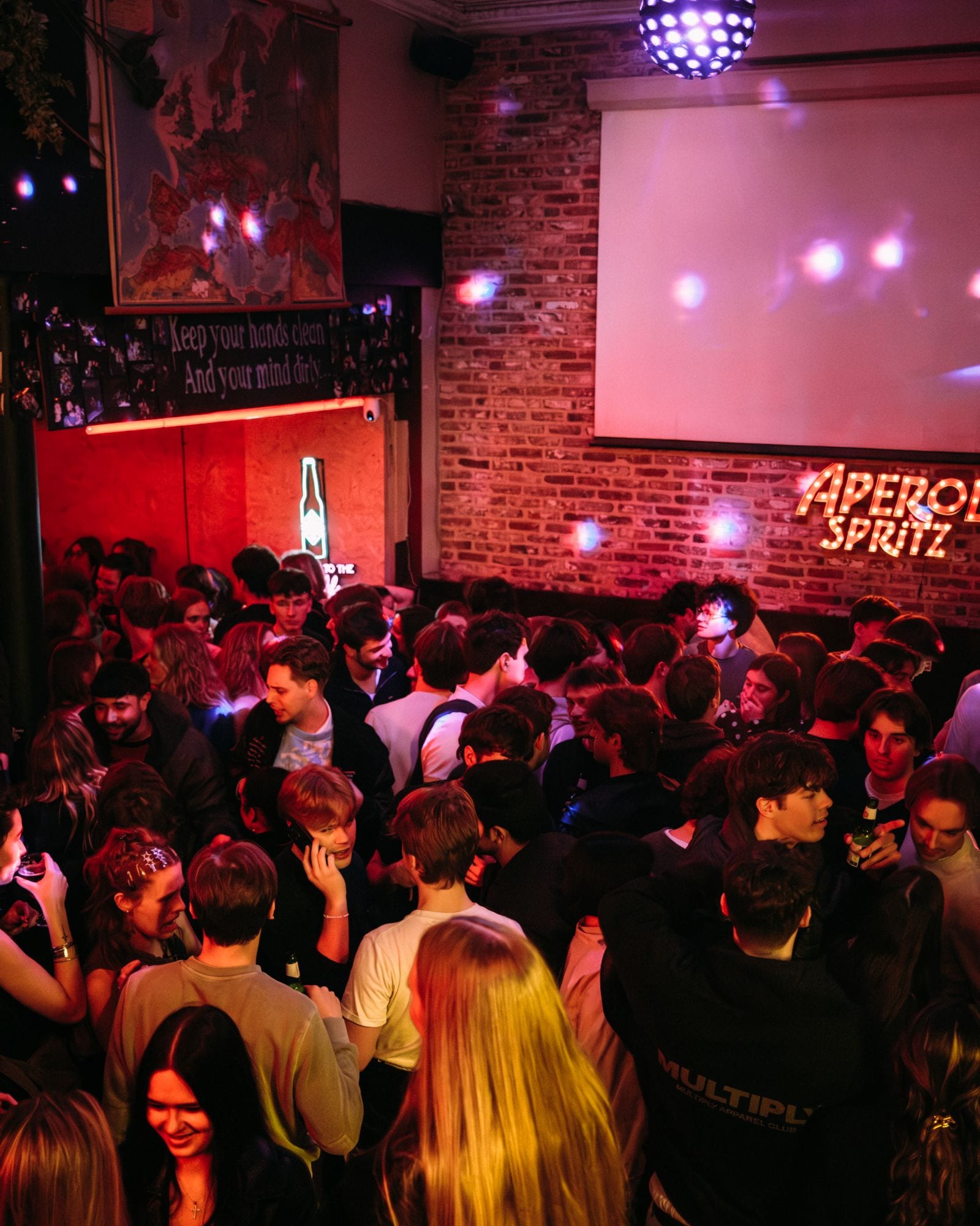 Een volle zaal onder een discobol, met een Aperol Spritz-bord tegen de bakstenen muur.